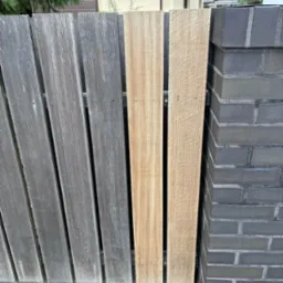 Odnowione drewniane ogrodzenie z desek o różnej kolorystyce, połączone z grafitowym, ceglanym słupkiem. Widoczne ślady renowacji i impregnacji drewna.