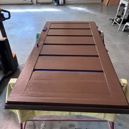 LASER SYSTEM KAMIL WIDAWSKI - Odnowione, brązowe drzwi z poziomymi panelami, leżące na stole warsztatowym. Widoczne detale wykończenia i faktura farby. W tle warsztat z narzędziami.