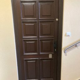 LASER SYSTEM KAMIL WIDAWSKI - Ciemnobrązowe, kasetonowe drzwi wejściowe z metalową klamką i wizjerem, zamontowane w jasnej ścianie klatki schodowej z białą listwą przypodłogową.