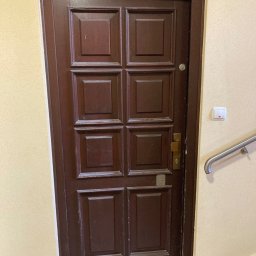 LASER SYSTEM KAMIL WIDAWSKI - Ciemnobrązowe, drewniane drzwi z kasetonami w beżowej ścianie klatki schodowej, widoczny fragment poręczy i przycisk dzwonka.