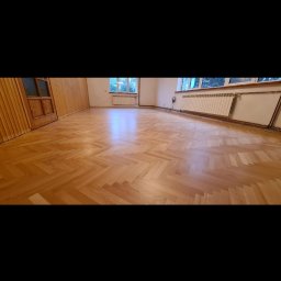 LASER SYSTEM KAMIL WIDAWSKI - Parkiet ułożony w jodełkę, po cyklinowaniu, z widocznym połyskiem. W tle okno i grzejnik. Naturalne odcienie drewna.