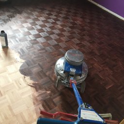 LASER SYSTEM KAMIL WIDAWSKI - Cyklinowanie parkietu w jodełkę. Połowa podłogi wycyklinowana, ukazując jaśniejszy odcień drewna. Widoczna maszyna do cyklinowania i butelka z preparatem.