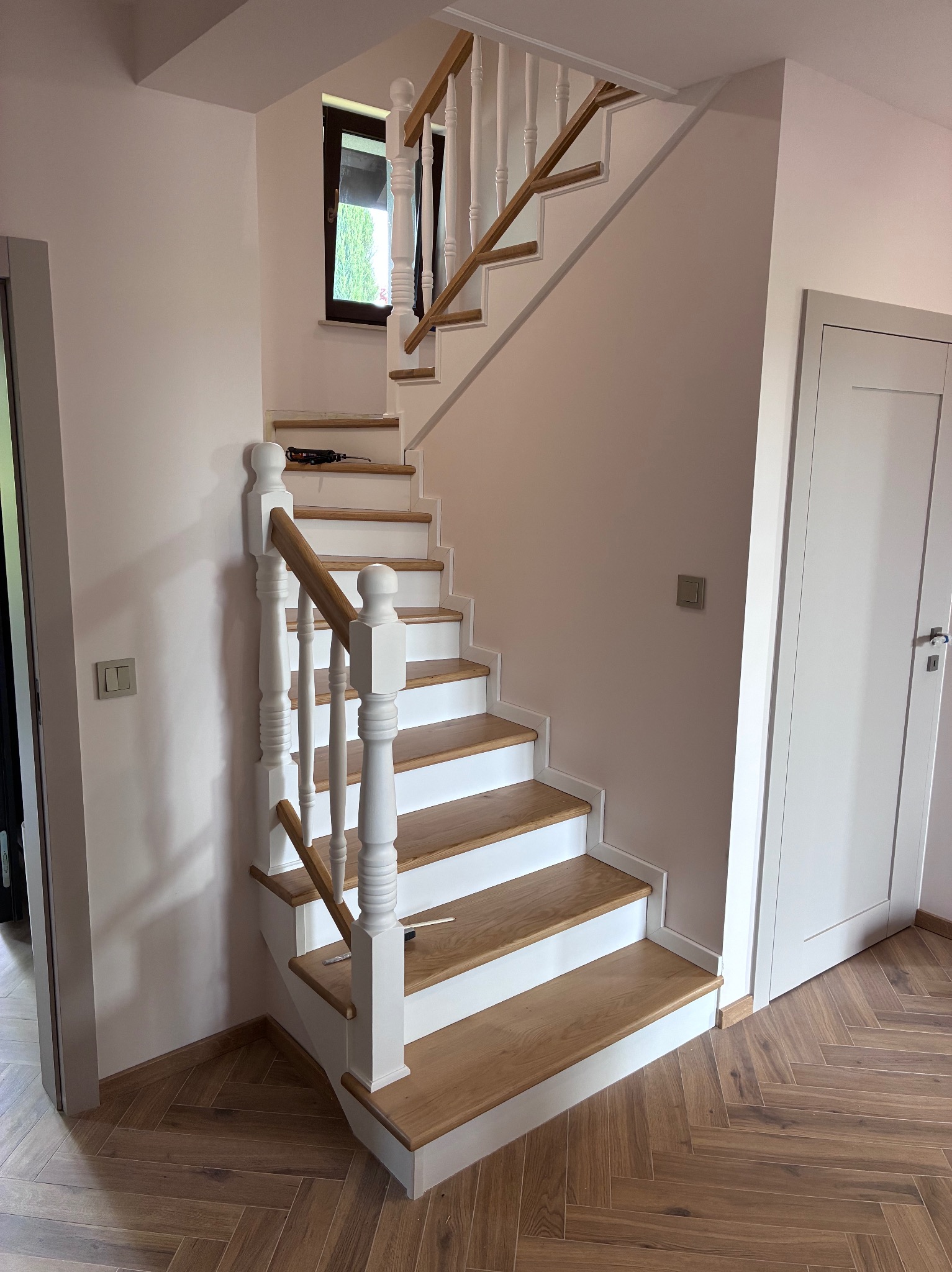 Jasne drewniane schody z białymi balustradami i stopniami, widoczne narzędzia na jednym ze stopni, podłoga ułożona w jodełkę, drzwi po bokach.