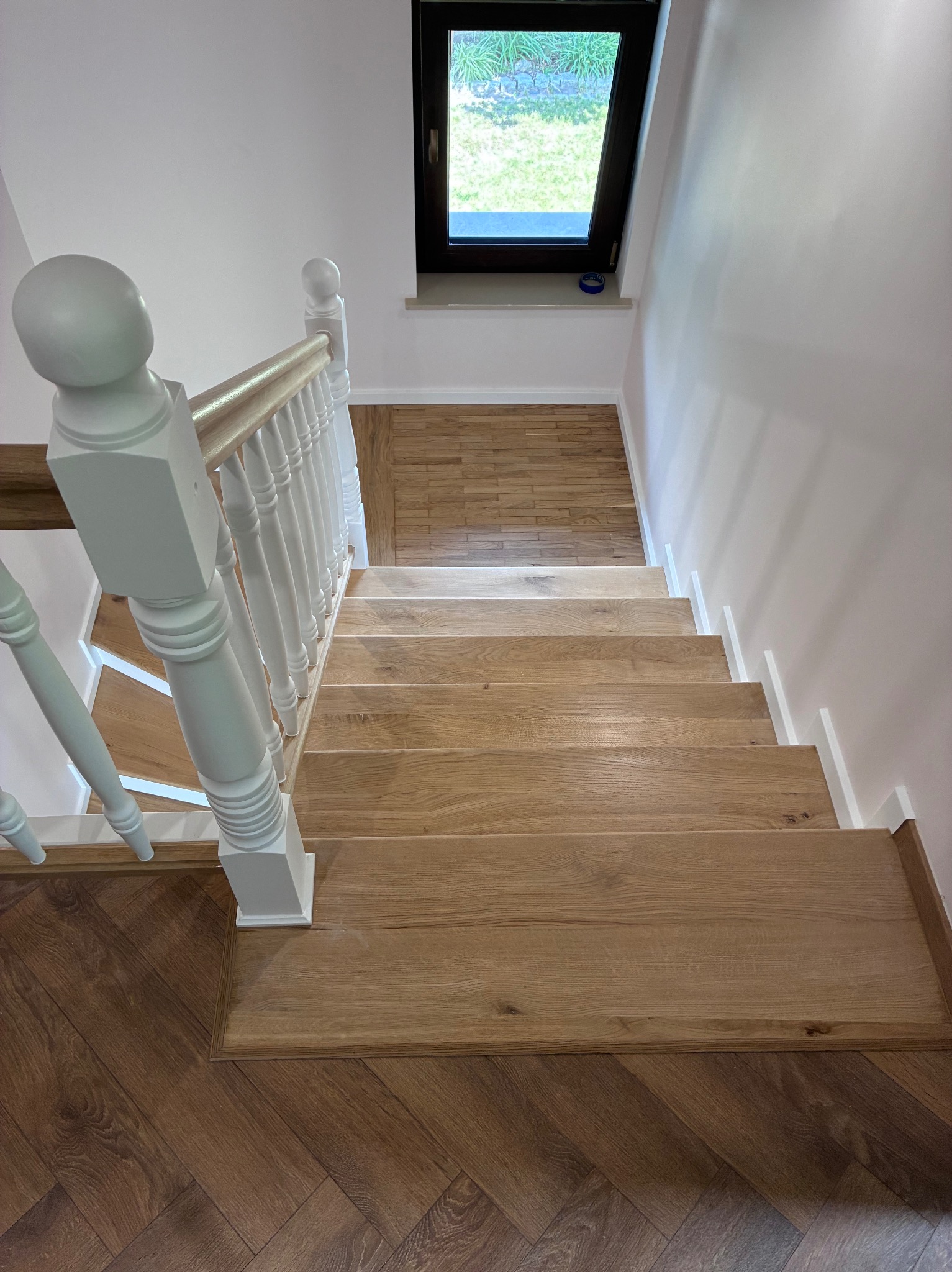 Drewniane schody z białymi balustradami, widok z góry, parkiet w jodełkę na podeście, okno dachowe w tle. Jasne, naturalne wykończenie drewna.