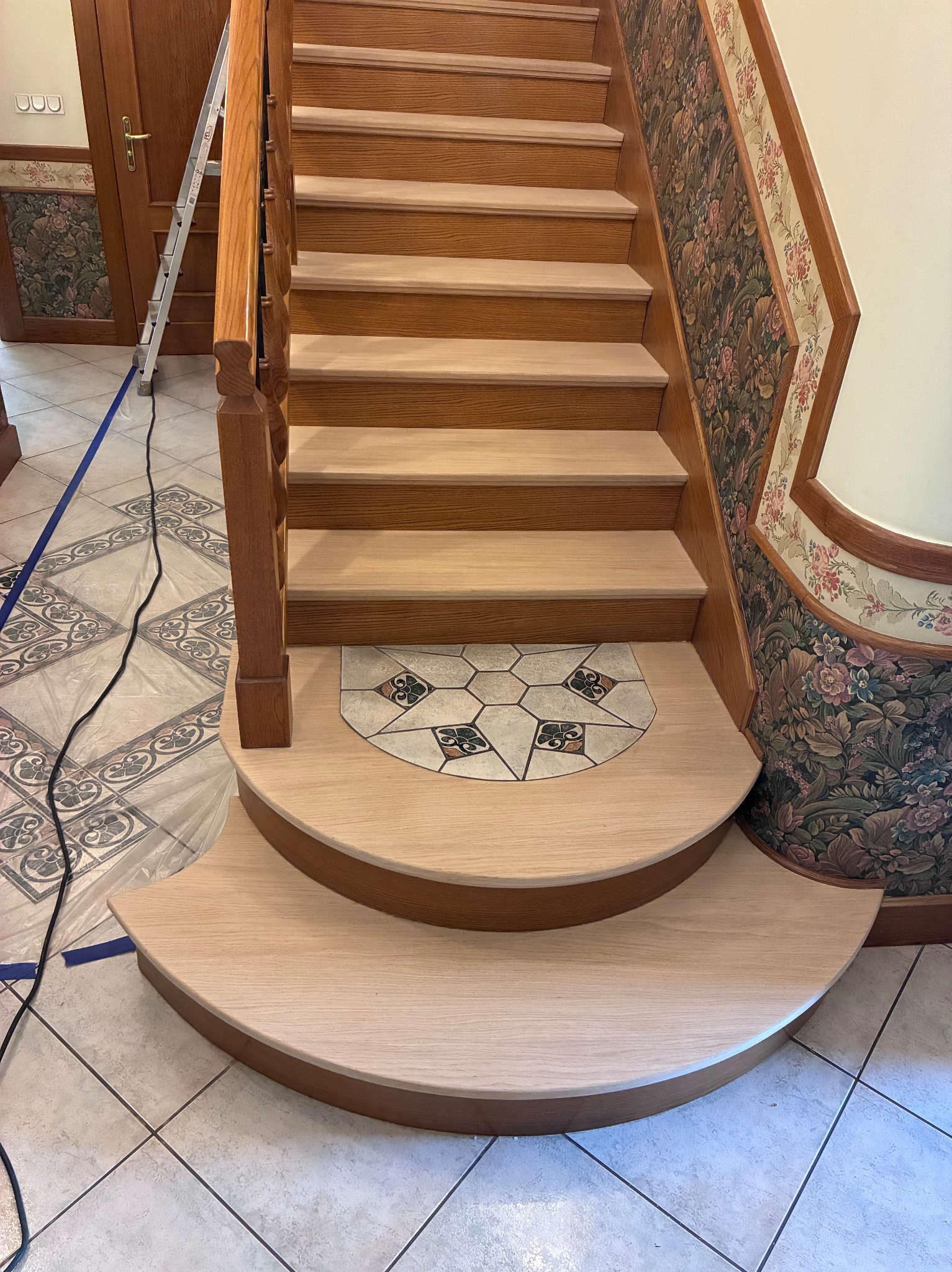 Jasne, odnowione drewniane schody z dekoracyjnymi elementami, zabezpieczone folią i taśmą malarską podczas prac wykończeniowych. Widoczna drabina w tle.