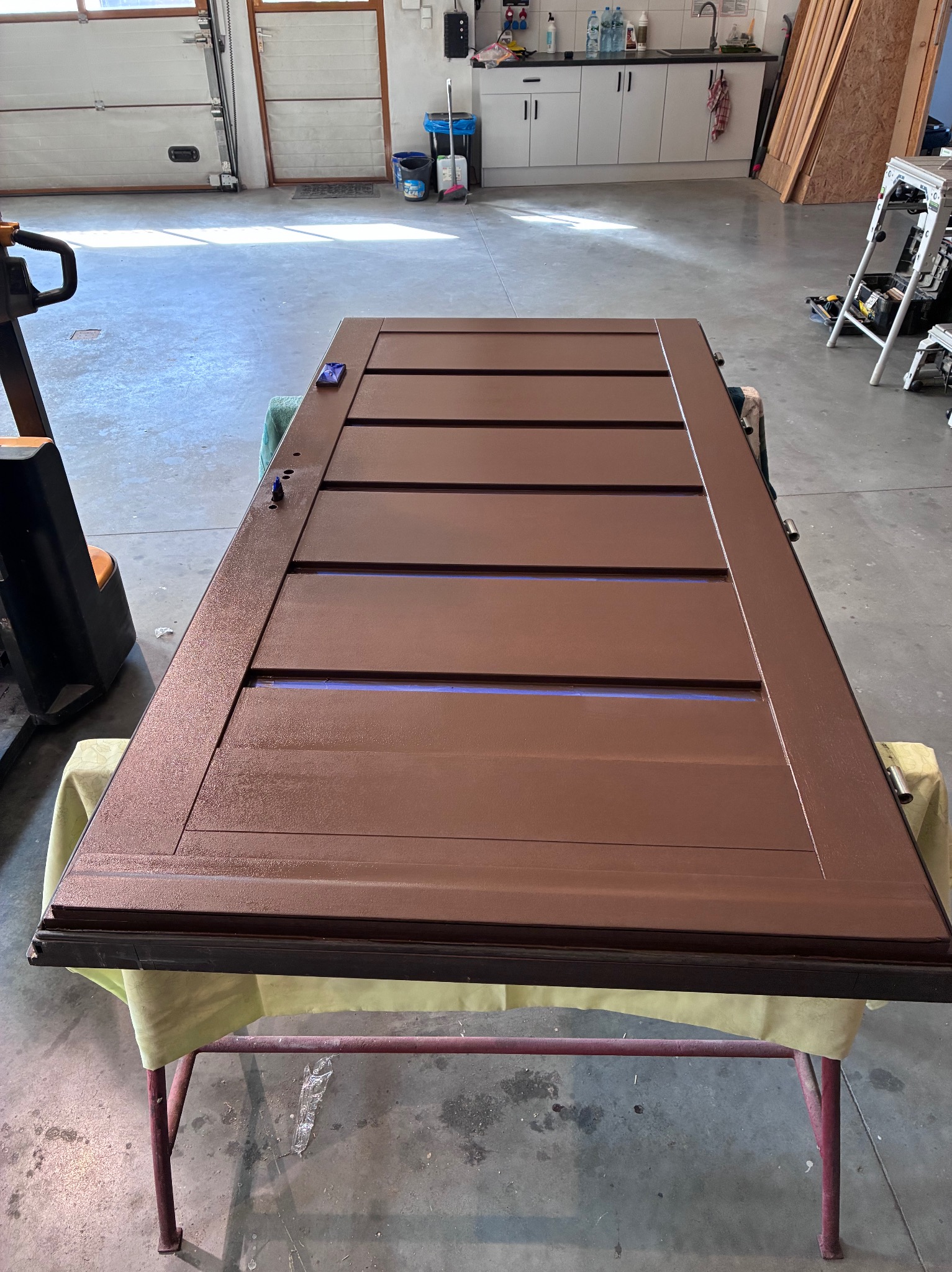 Odnowione, brązowe drzwi z poziomymi panelami, leżące na stole warsztatowym. Widoczne detale wykończenia i faktura farby. W tle warsztat z narzędziami.