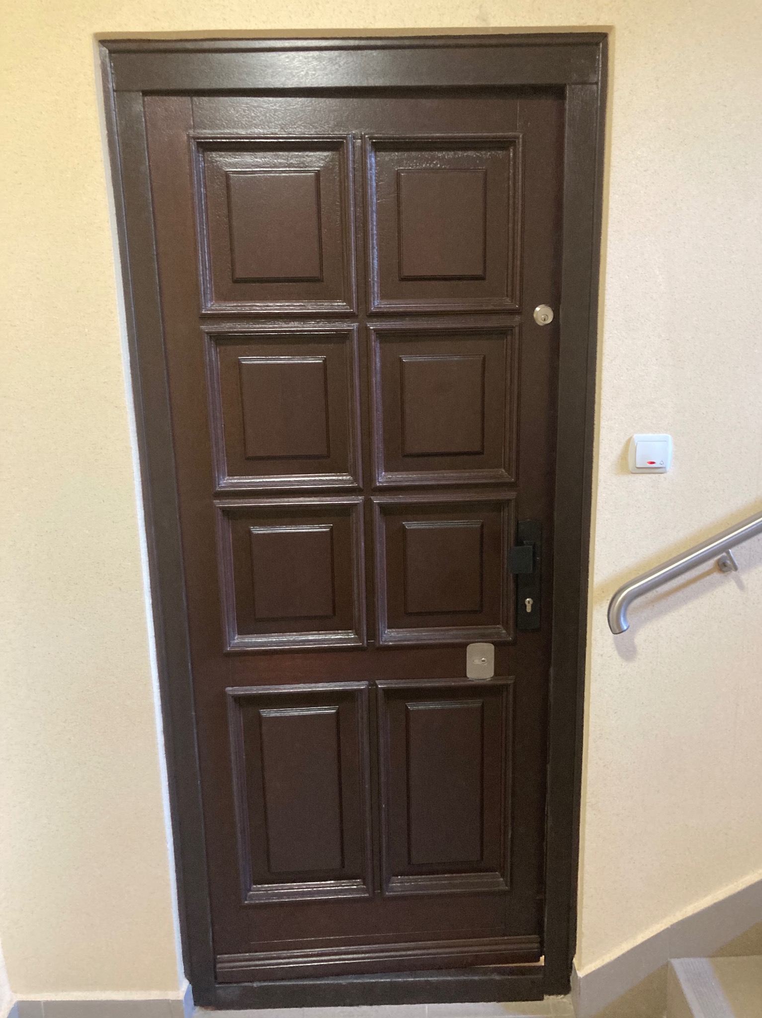 Ciemnobrązowe, kasetonowe drzwi wejściowe z metalową klamką i wizjerem, zamontowane w jasnej ścianie klatki schodowej z białą listwą przypodłogową.