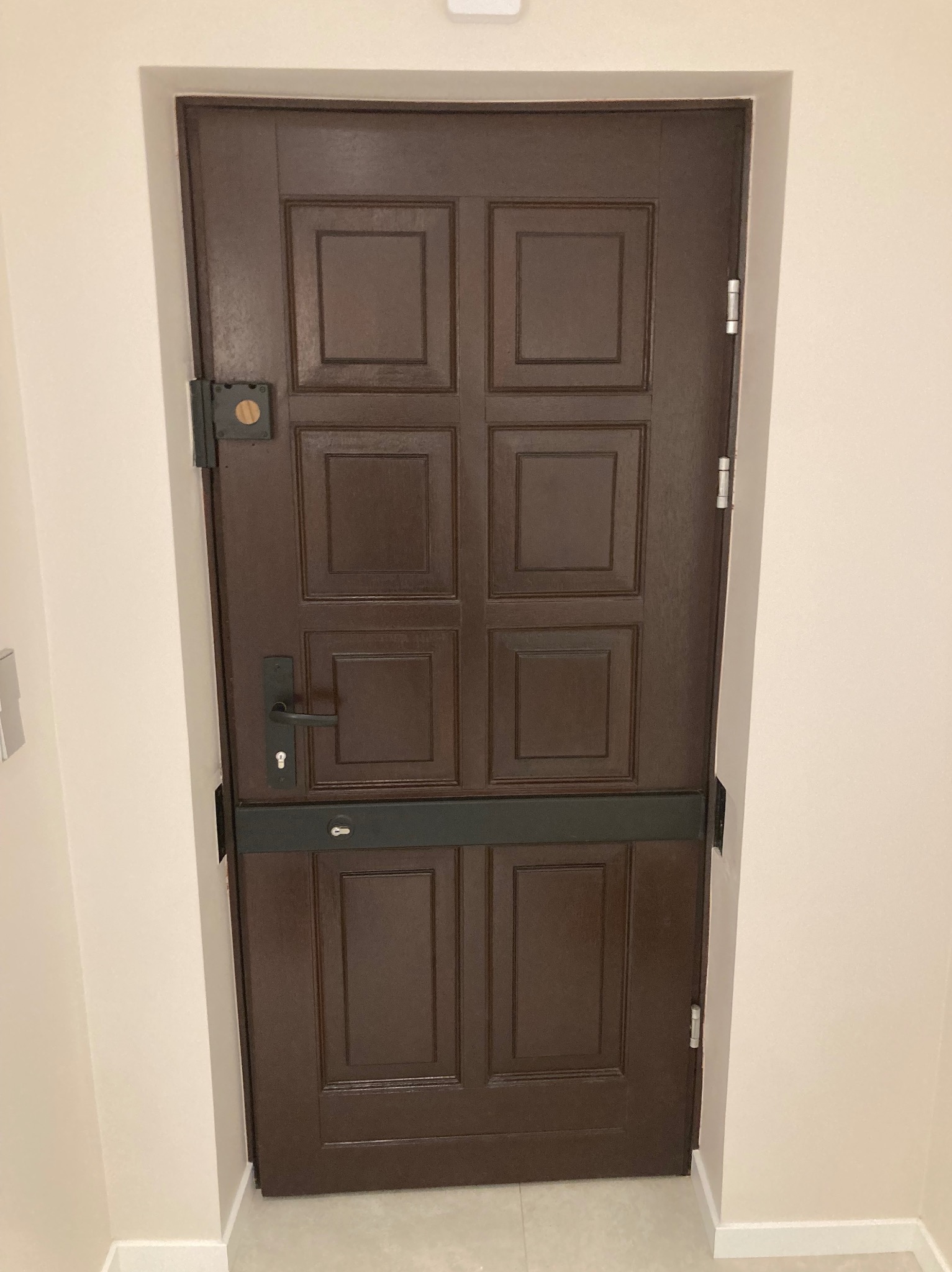 Ciemnobrązowe, drewniane drzwi wewnętrzne z metalowymi okuciami i czarną poprzeczką zabezpieczającą, widoczne na tle jasnej ściany.