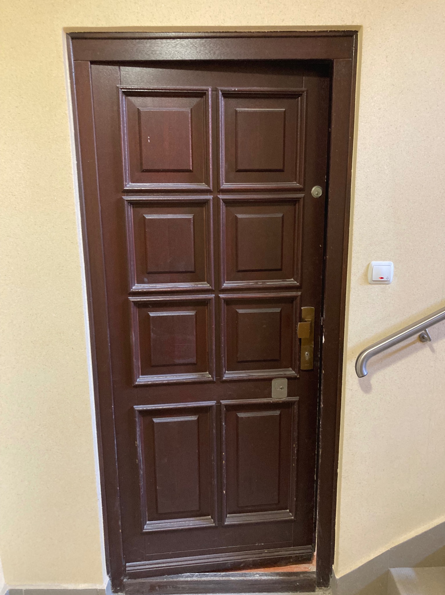 Ciemnobrązowe, drewniane drzwi z kasetonami w beżowej ścianie klatki schodowej, widoczny fragment poręczy i przycisk dzwonka.