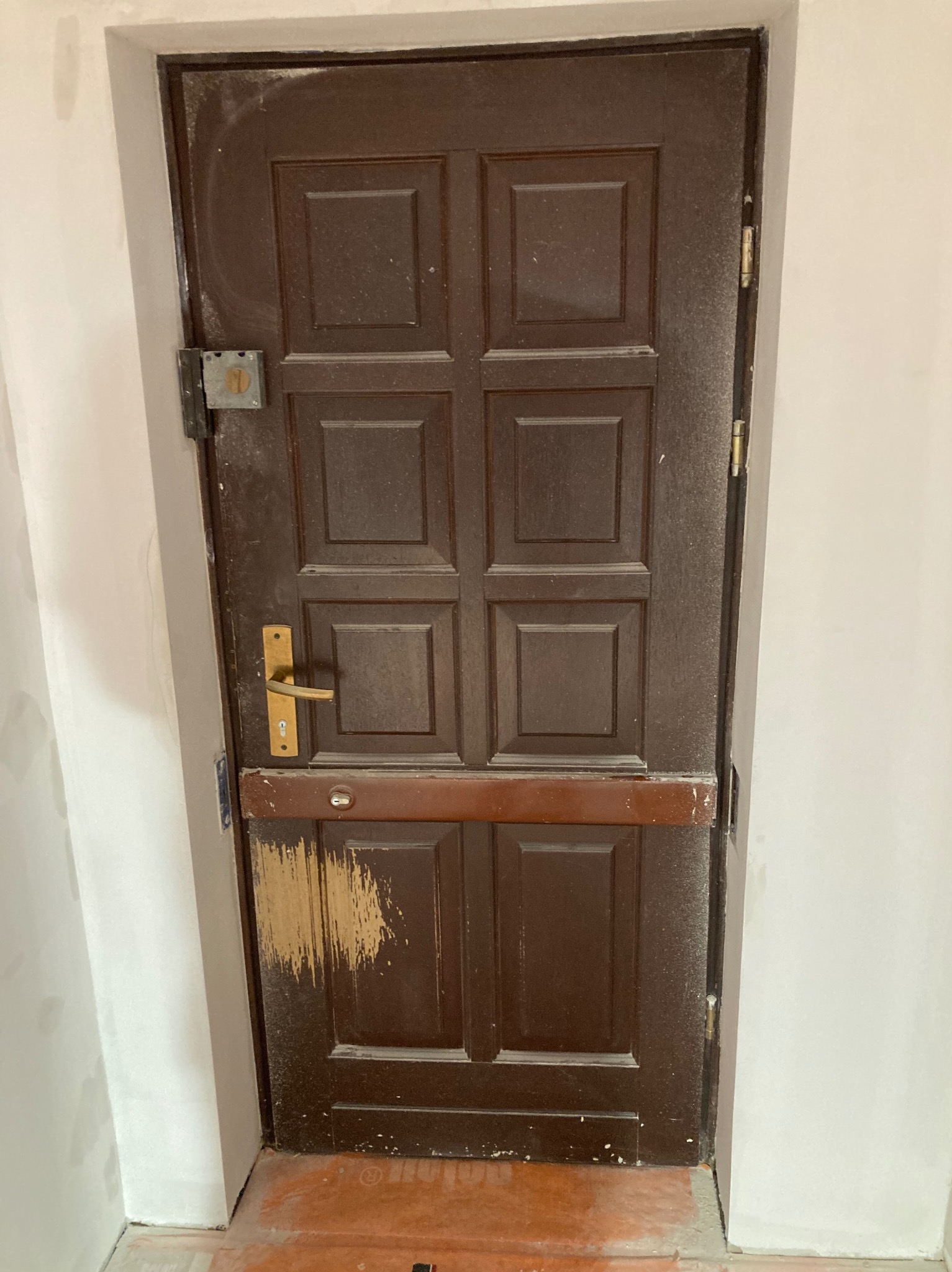 Stare, brązowe drzwi wymagające renowacji z widocznymi śladami uszkodzeń i odchodzącą farbą w dolnej części. Zauważalny kurz i zabrudzenia na całej powierzchni.