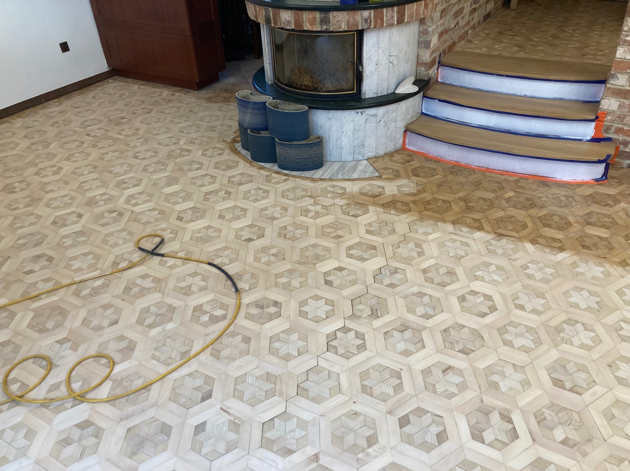 Parkiet z wzorem gwiazdy w heksagonach, częściowo odnowiony, z widocznymi materiałami ściernymi obok kominka i zabezpieczonymi schodami. Widoczny kabel od maszyny.