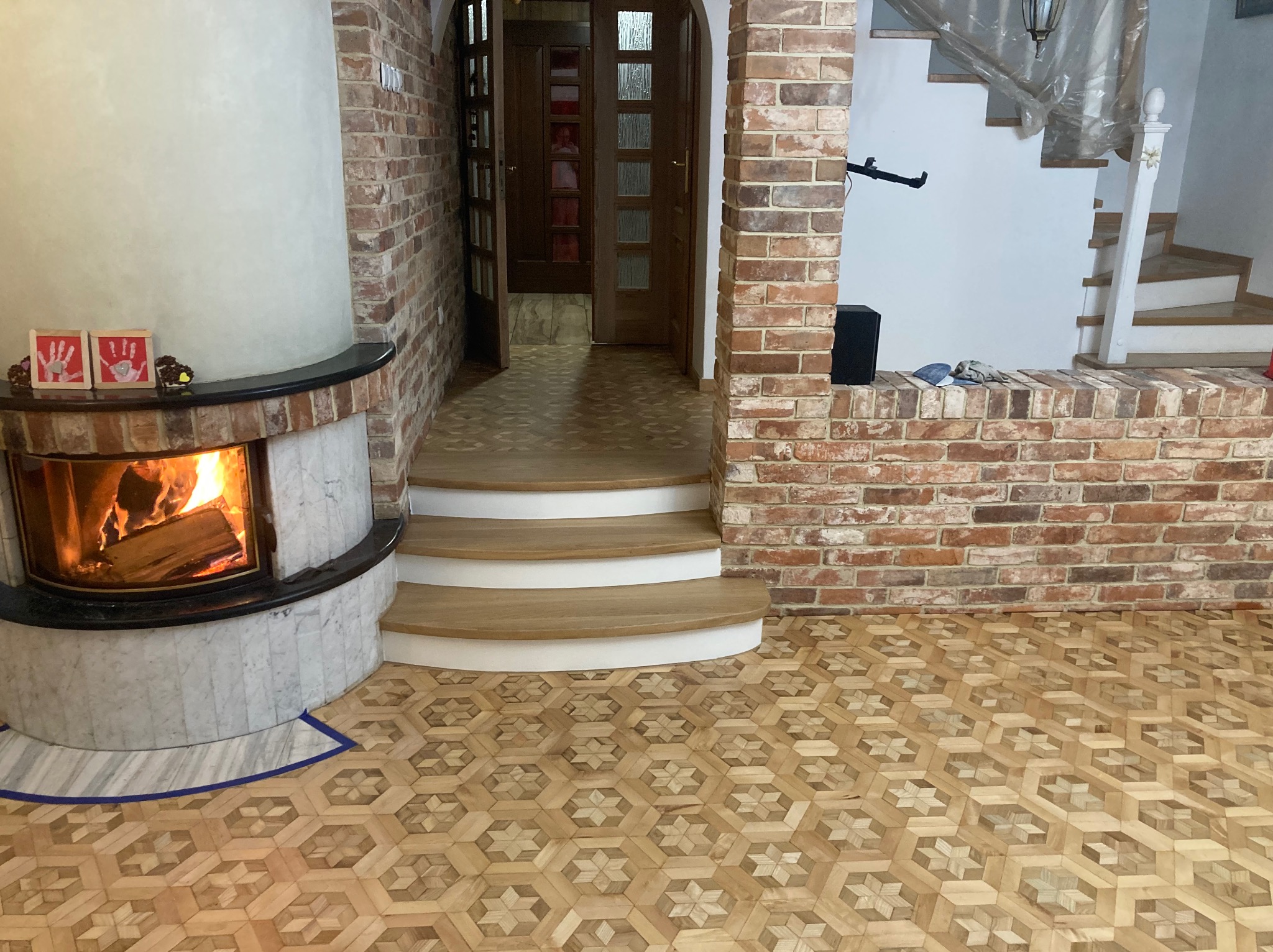 Salon z parkietem w geometryczny wzór, kominkiem z paleniskiem i ceglanymi elementami dekoracyjnymi. Widoczne drewniane schody i otwarte drzwi.