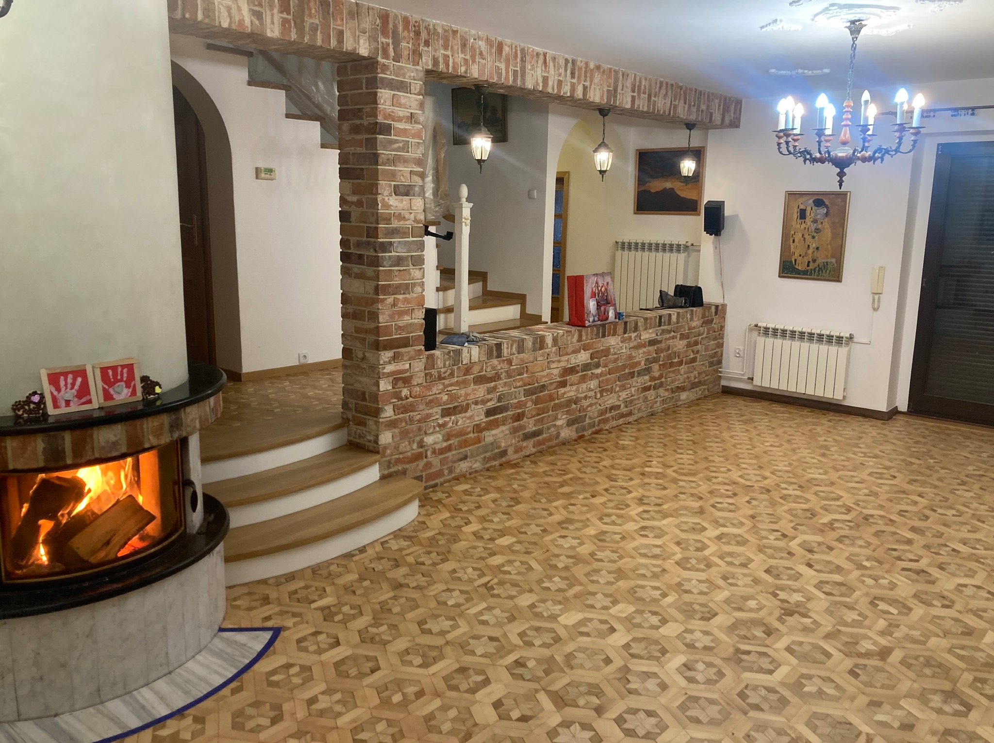 Parkiet w geometryczny wzór, kominek z paleniskiem, ceglana ściana i schody w przestronnym wnętrzu. Żyrandol dodaje elegancji.