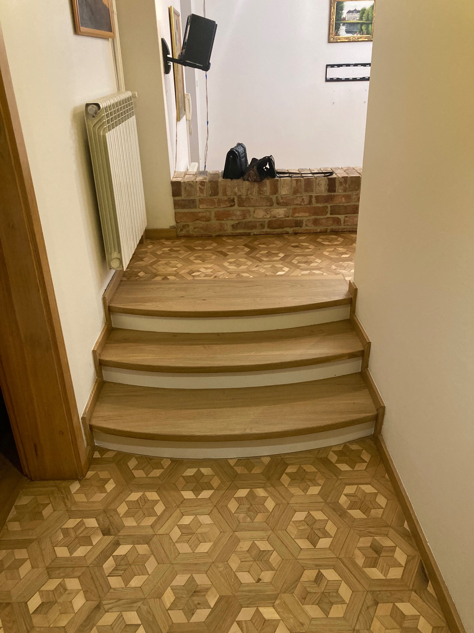 Drewniane schody z parkietem o wzorze geometrycznym, widok z góry, ciepłe oświetlenie. Widoczny fragment grzejnika i ceglanej ściany w tle.