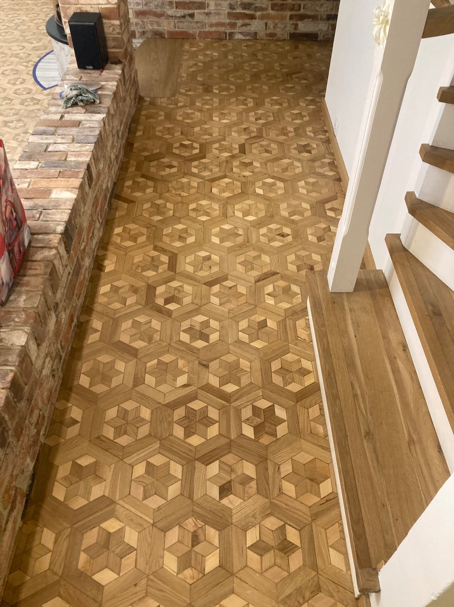 Ukończona podłoga z parkietu o wzorze geometrycznym, widoczna z perspektywy góry, sąsiadująca z ceglaną ścianą i drewnianymi schodami.