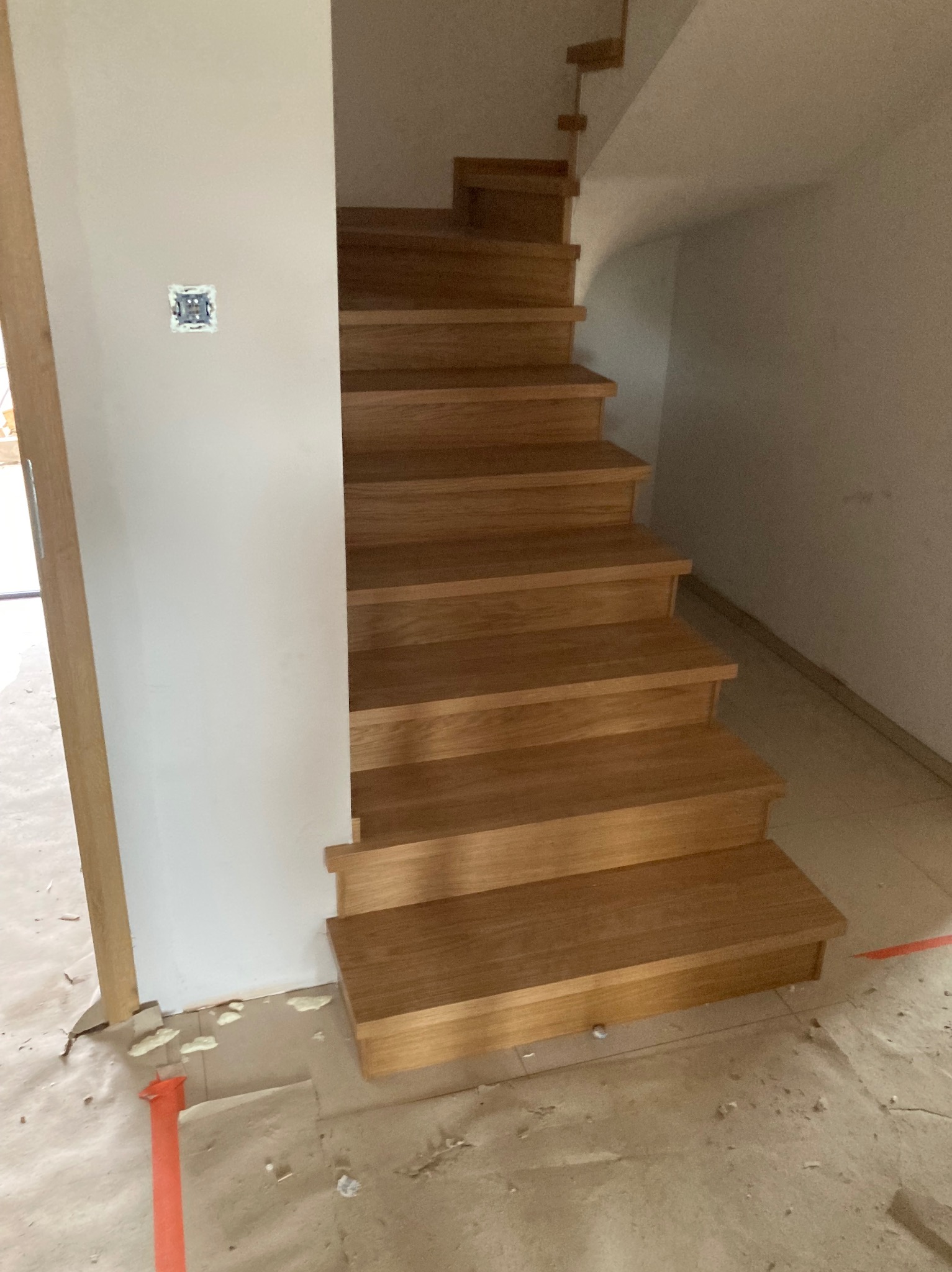 Dębowe schody z widocznym fragmentem ściany z gniazdkiem elektrycznym. Schody w jasnym odcieniu drewna, prowadzące w górę, w trakcie montażu, widoczne zabezpieczenia podłogi.