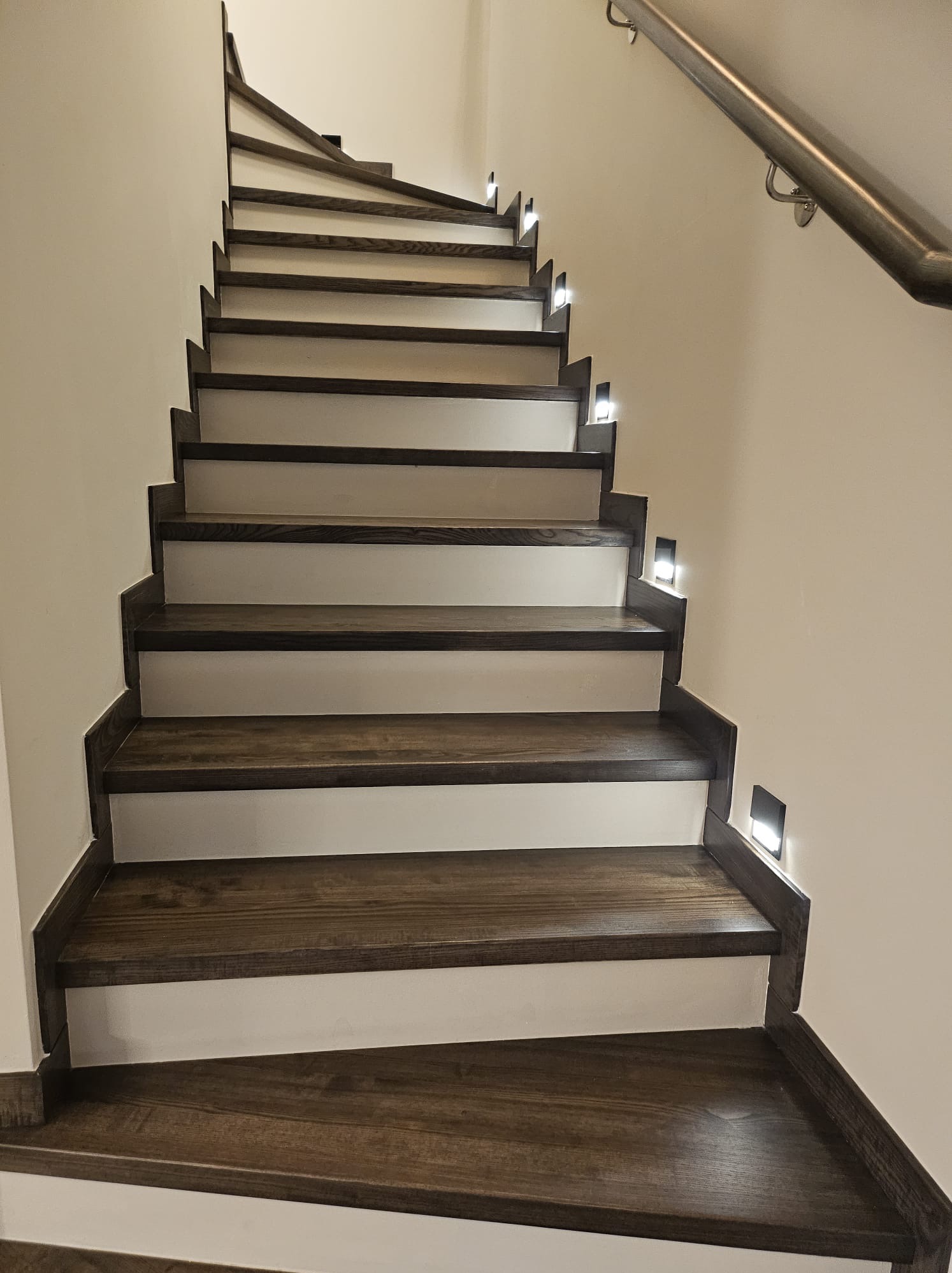 Drewniane schody z białymi podstopnicami i oświetleniem punktowym w ścianie, widok z dołu na górę, poręcz ze stali nierdzewnej po prawej stronie.