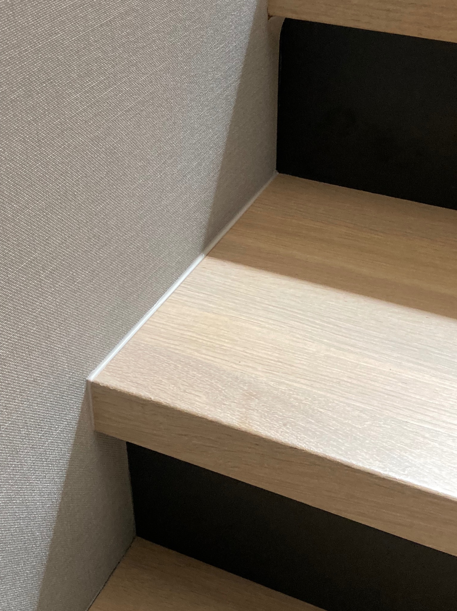 Jasne drewniane schody z czarnymi podstopnicami i szarą ścianą. Minimalistyczny design, oświetlenie z góry podkreśla teksturę drewna.