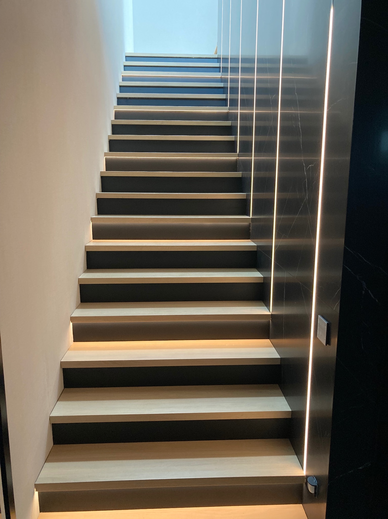 Nowoczesne schody z podświetleniem LED, jasne drewno na stopniach, czarne podstopnice i marmurowe wykończenie ściany z pionowymi paskami LED.