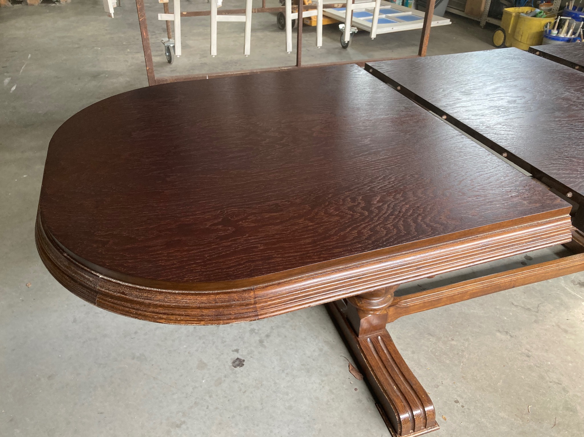 Odnowiony, rozkładany stół z ciemnego drewna dębowego, z widocznym rysunkiem słojów, na tle warsztatu. Stół po renowacji blatu.