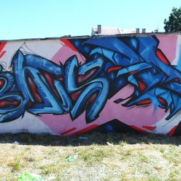 Piotr Wolski - Kolorowe graffiti z niebieskimi i czerwonymi akcentami na ścianie z różowym tłem, widoczne trawa i fragment chodnika. Street art w plenerze.