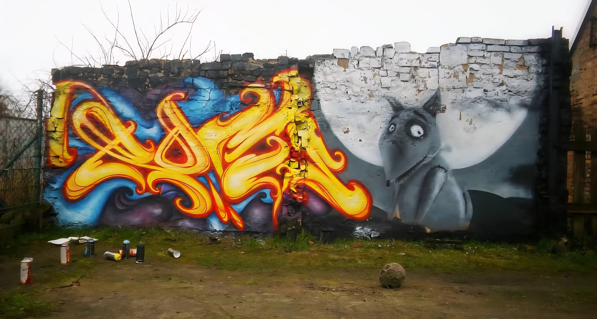 Mur z graffiti: abstrakcyjne, płomienne wzory w żółcieni i błękicie oraz postać zwierzęcia na tle księżyca. Puszki z farbą leżą na ziemi.