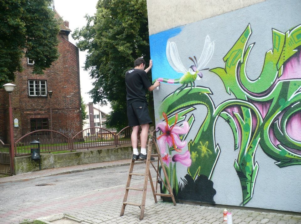 Artysta na drabinie tworzy mural ze stylizowaną ważką i kwiatami na szarej ścianie. W tle budynek z czerwonej cegły i zieleń drzew.