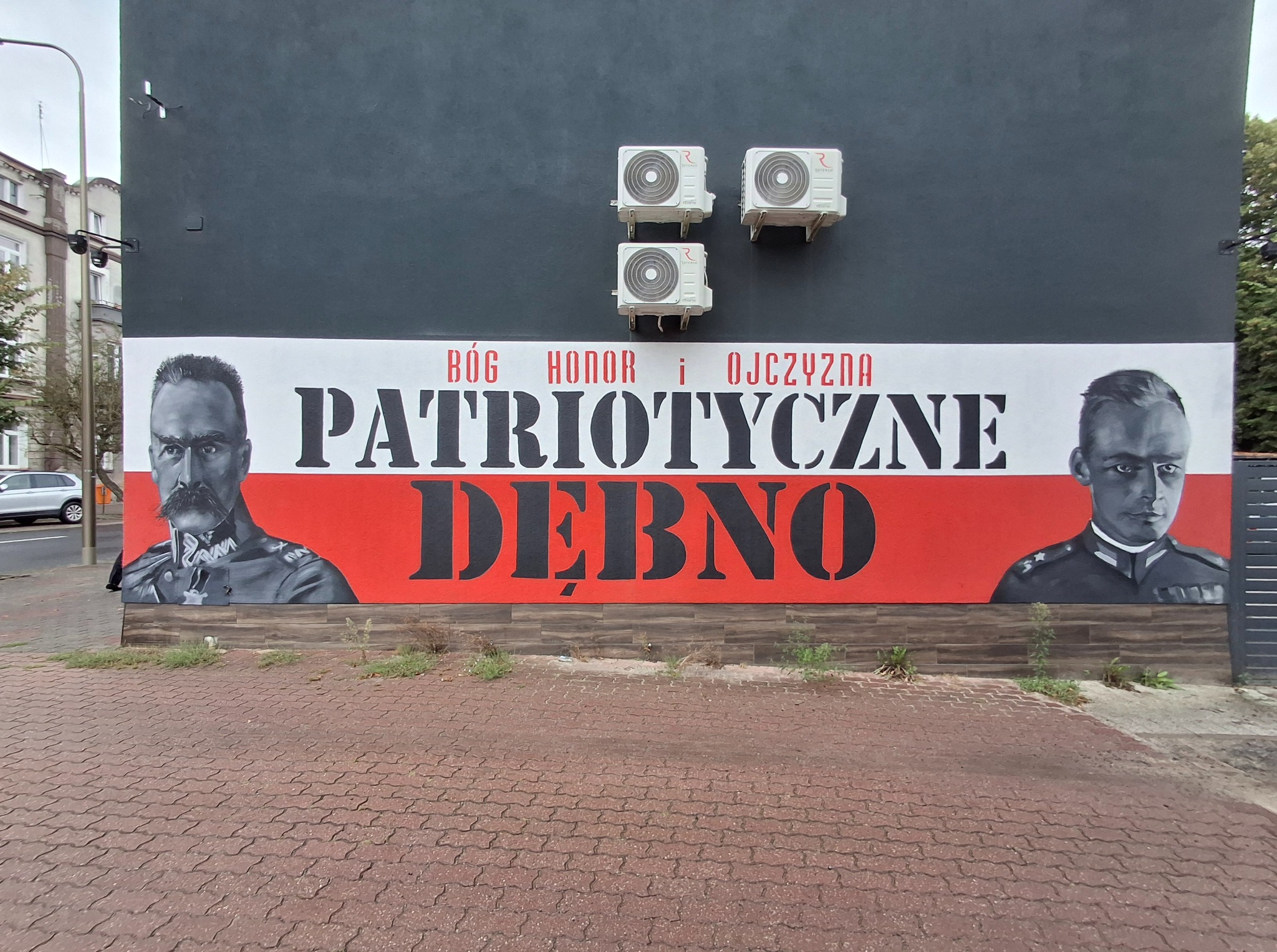 Mural na szarej ścianie budynku z hasłem 'Bóg Honor i Ojczyzna' oraz wizerunkami dwóch postaci w mundurach. Dębno.