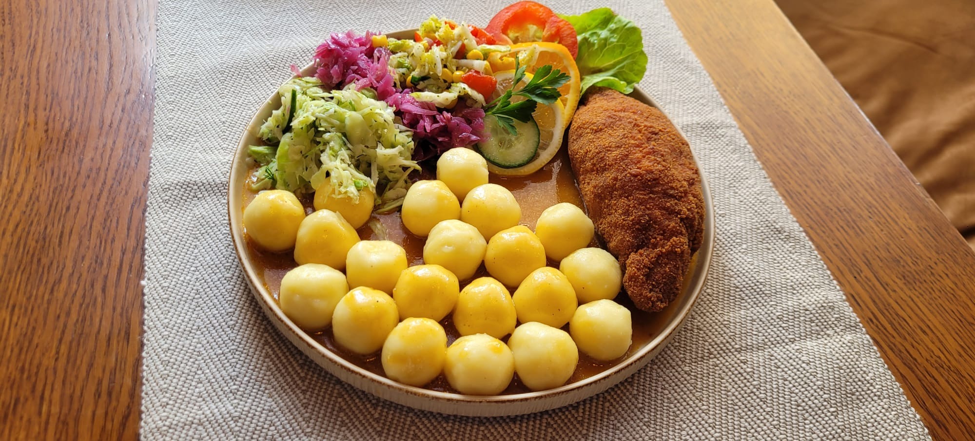 Apetyczna kompozycja na talerzu: kluski śląskie w sosie, kotlet schabowy w panierce i kolorowe surówki warzywne z dodatkiem ziół, na lnianym obrusie.