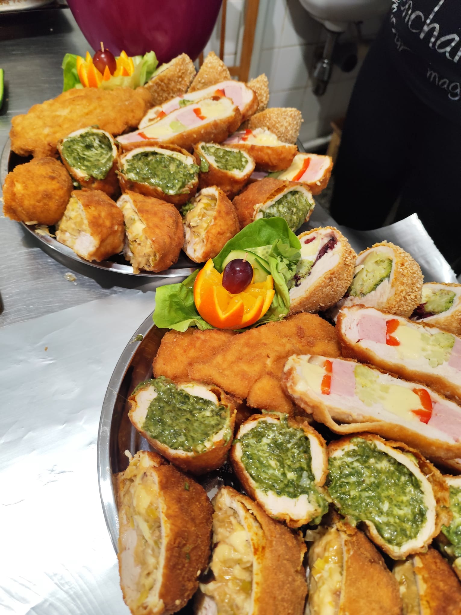 Dwa metalowe półmiski z przekąskami cateringowymi: roladki z szynką i serem, kotlety ze szpinakiem i kulki mięsne, ozdobione pomarańczą i sałatą.