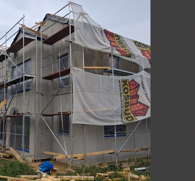 Dom w trakcie tynkowania, osłonięty siatką z logo, z widocznymi rusztowaniami i przygotowanymi oknami. Prace na zewnątrz budynku w pochmurny dzień.