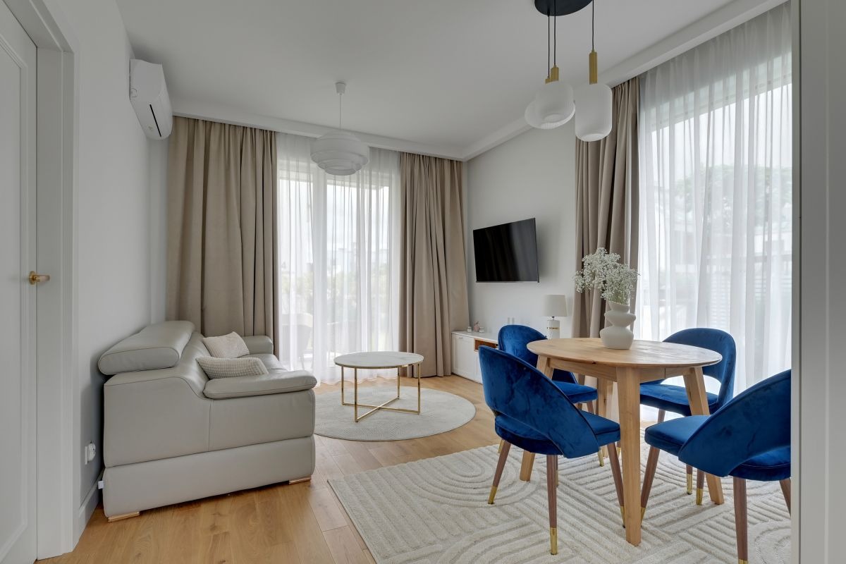 Jasne wnętrze: szara sofa, okrągły stół z niebieskimi krzesłami, beżowe zasłony w oknach, drewniana podłoga, minimalistyczny wystrój. Nowoczesne mieszkanie.