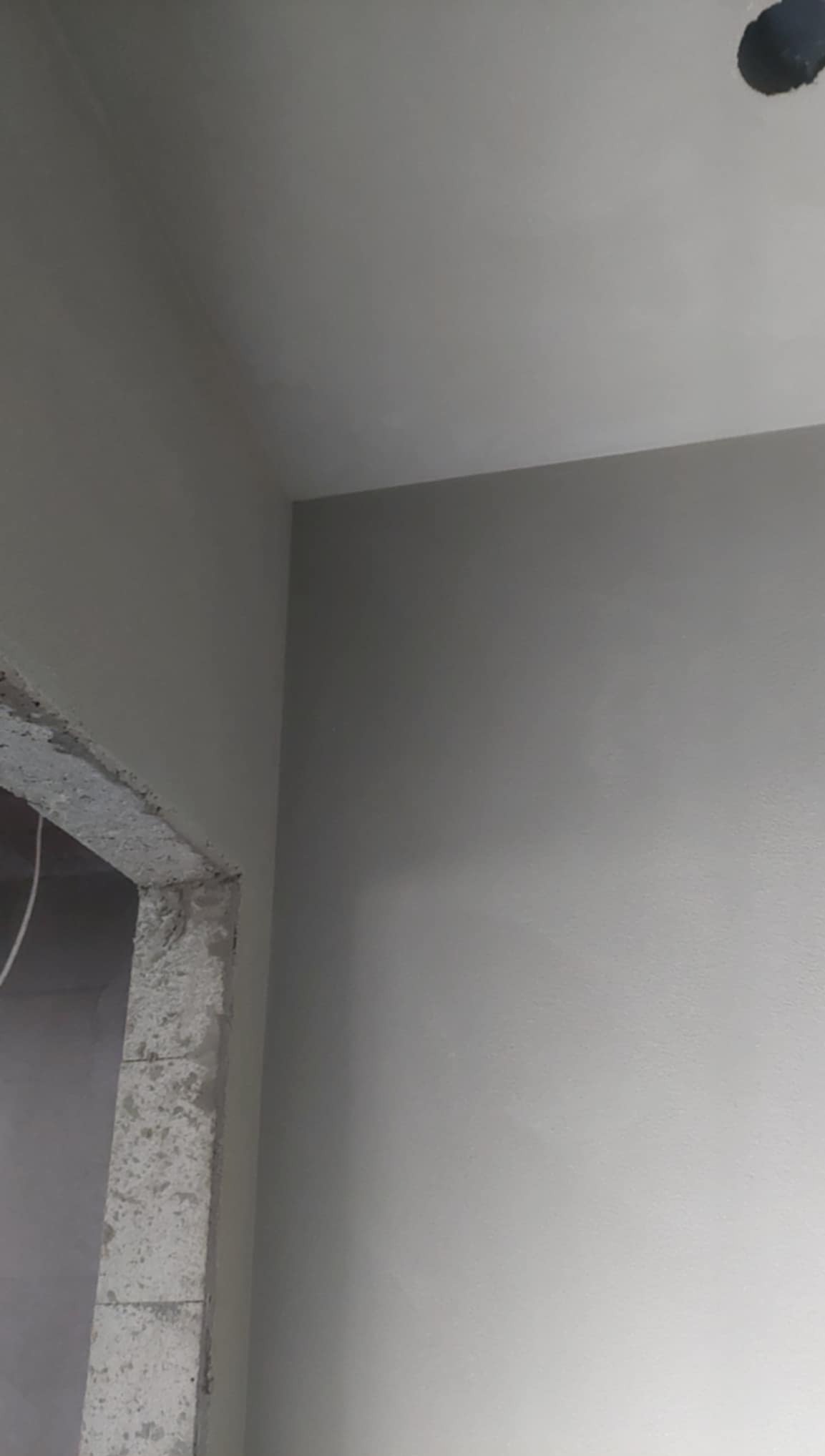 Gładka, szara ściana po tynkowaniu maszynowym, widoczny fragment surowego ościeża drzwiowego. Minimalistyczne ujęcie detalu wykończenia wnętrza.