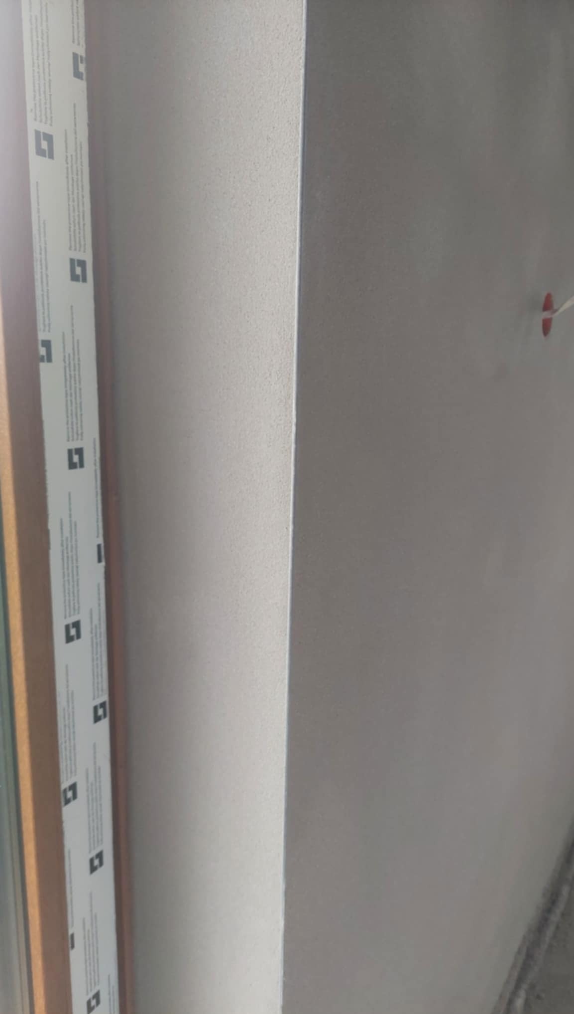 Gładka, szara ściana po tynkowaniu maszynowym, widoczna obok ramy okiennej z białymi oznaczeniami. Minimalistyczne ujęcie wykończonej powierzchni.