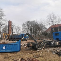 VOS Rafał Osadnik - Demontaż ceglanego komina na tle gruzu i drzew. Widoczne ciężarówki i kontener, sugerujące prace rozbiórkowe i wywóz gruzu. Żółta koparka w tle.