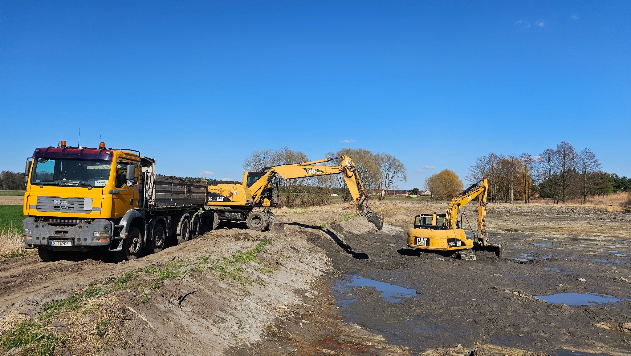 Żółty zestaw maszyn budowlanych: ciężarówka MAN i dwie koparki CAT, wykonujące prace ziemne w błotnistym terenie pod błękitnym niebem.