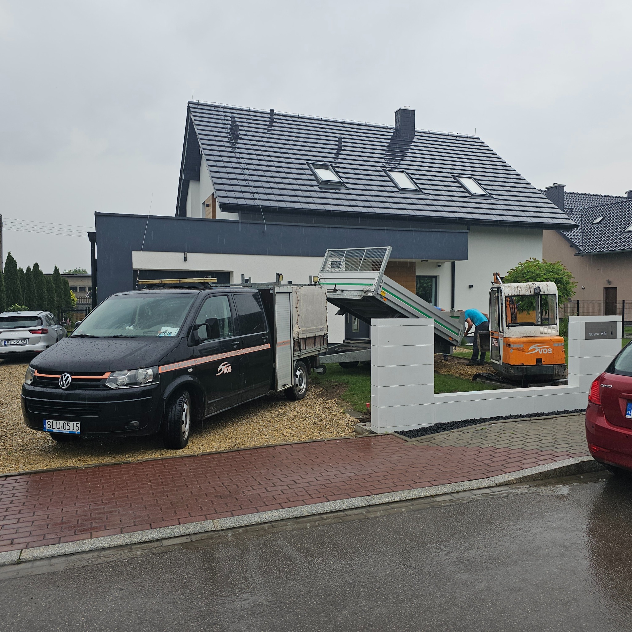 Ciężarówka i minikoparka VOS na żwirowym podjeździe, pracownik w niebieskiej koszulce przyczepia przyczepkę. Nowoczesny dom z szarą dachówką w tle, pochmurny dzień.