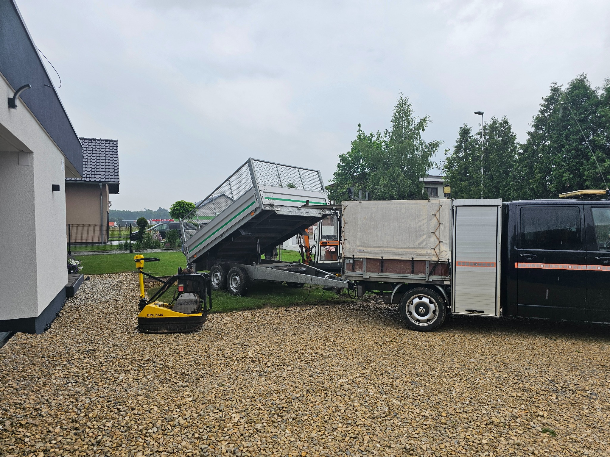 Podniesiona przyczepa, ubijarka wibracyjna i samochód na żwirowym podłożu. Sprzęt gotowy do utwardzania terenu przy domu.