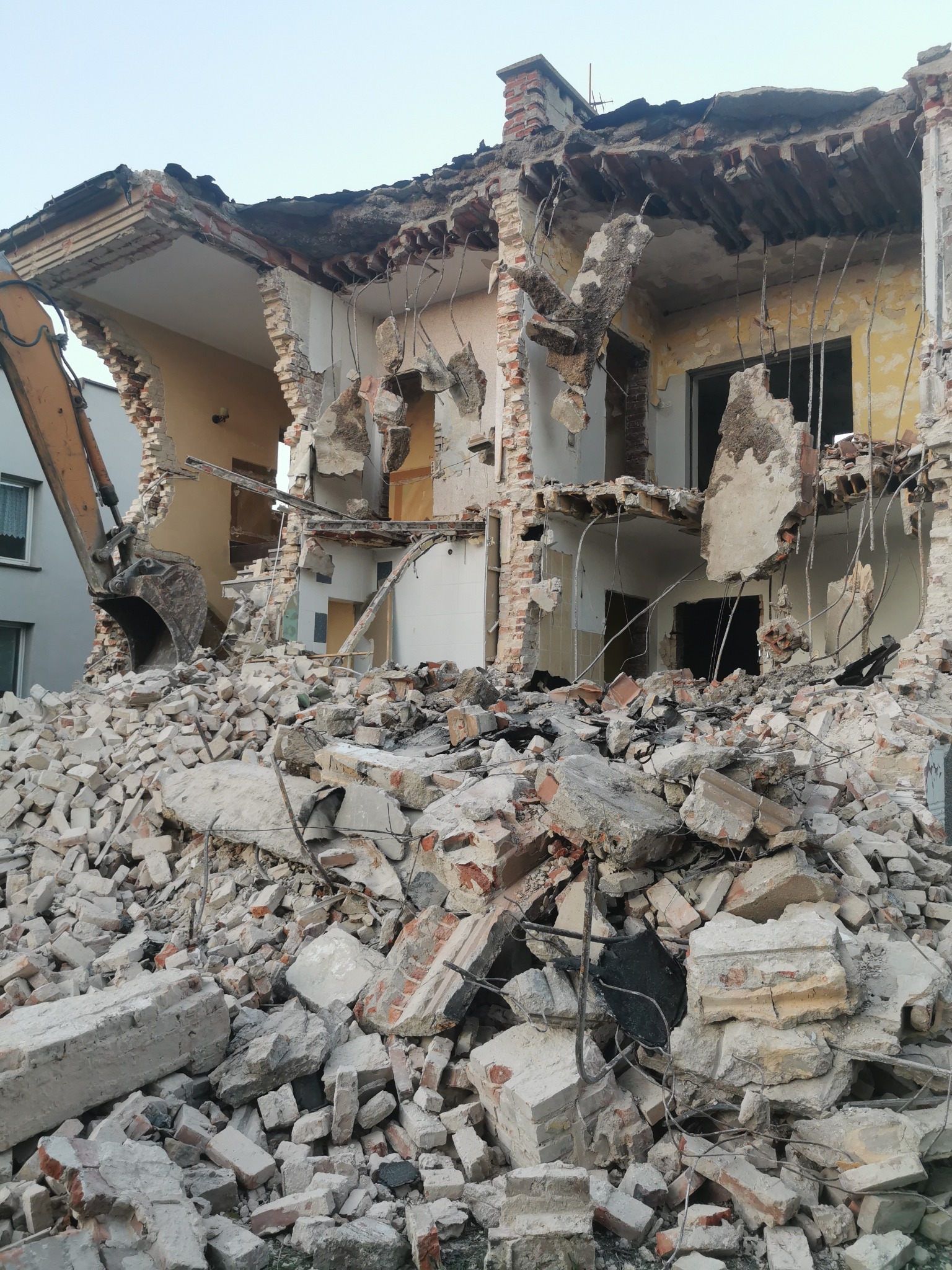 Częściowo zburzony budynek z widoczną koparką i stertą gruzu, ukazujący postęp rozbiórki. Odsłonięte wnętrza pomieszczeń.