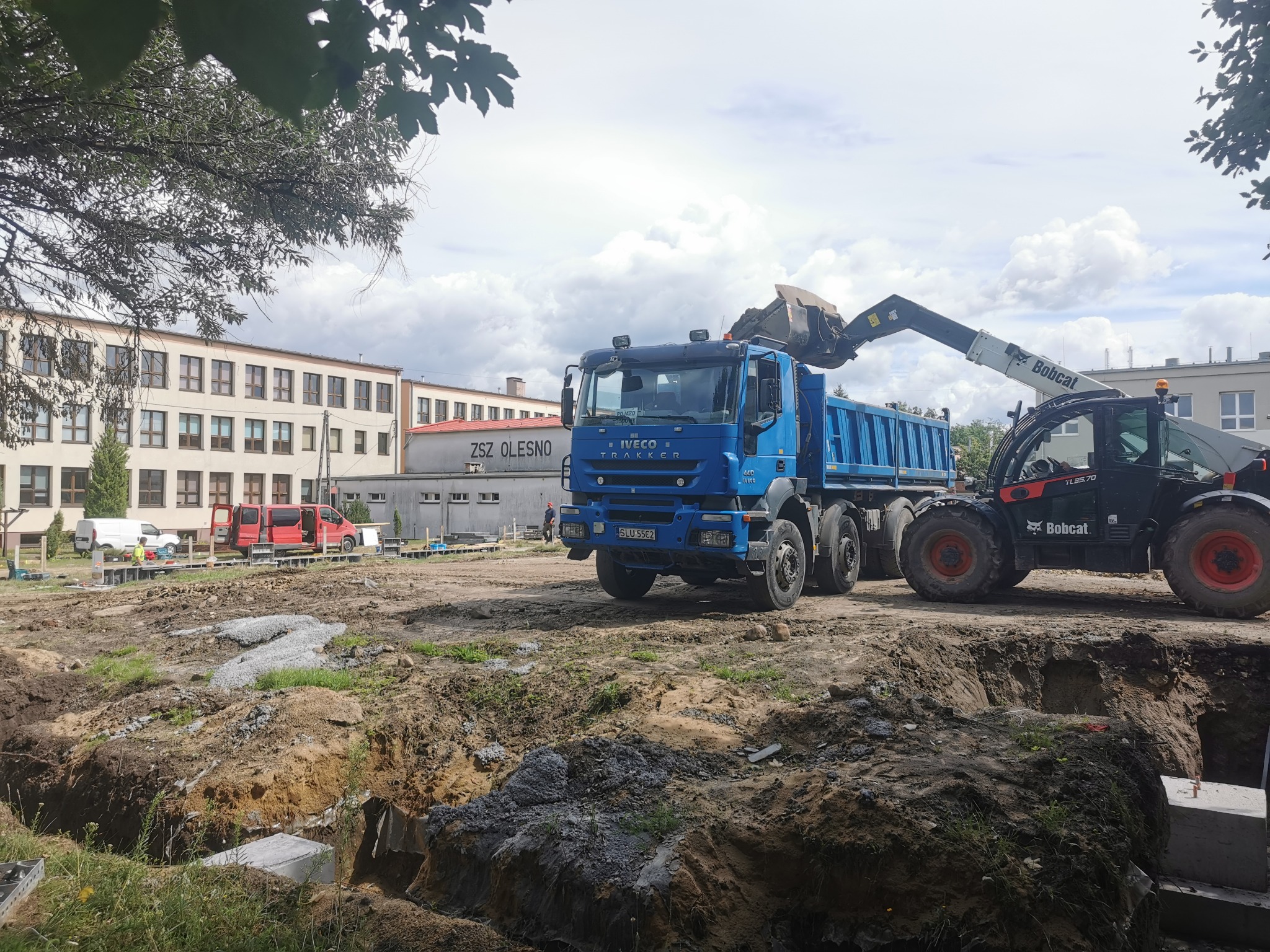 Niebieska ciężarówka Iveco i ładowarka Bobcat na placu budowy. Widoczne wykopy pod fundamenty i budynek w tle. Praca w toku.