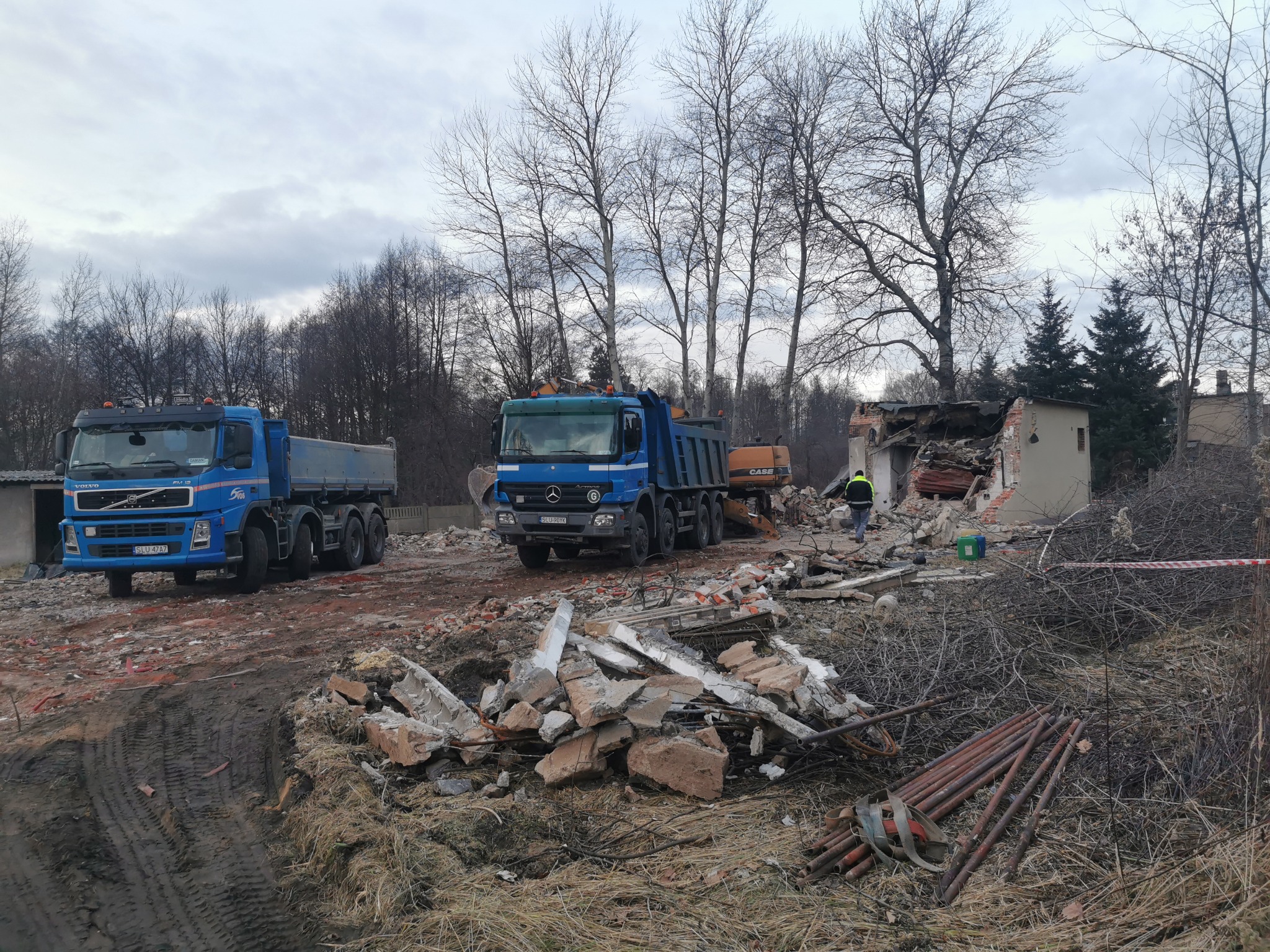 Dwa niebieskie samochody ciężarowe i koparka na terenie rozbiórki budynku z gruzem. Jeden pracownik w żółtej kamizelce. Pochmurne niebo.