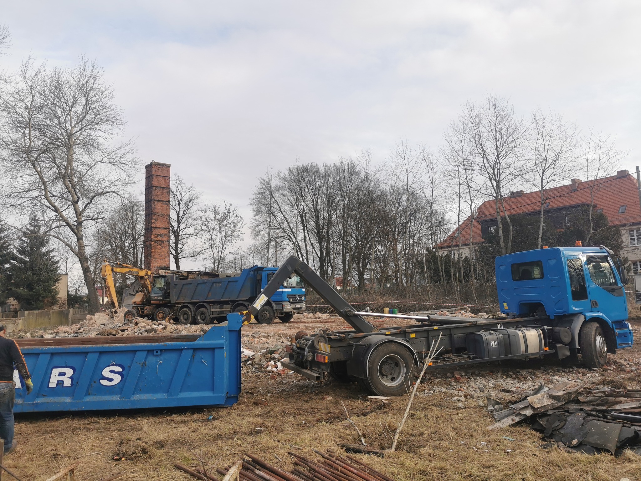 Demontaż ceglanego komina na tle gruzu i drzew. Widoczne ciężarówki i kontener, sugerujące prace rozbiórkowe i wywóz gruzu. Żółta koparka w tle.