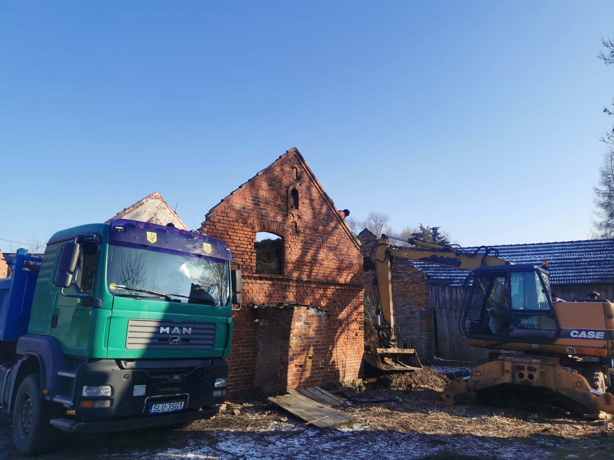 Rozbiórka ceglanego budynku z użyciem ciężkiego sprzętu: zielonej ciężarówki MAN i pomarańczowej koparki CASE 20, w słoneczny dzień.