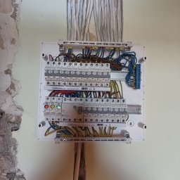 Mariusz Skonieczny - Rozdzielnica elektryczna z licznymi bezpiecznikami i przewodami w trakcie montażu na ścianie z widocznymi śladami prac remontowych.