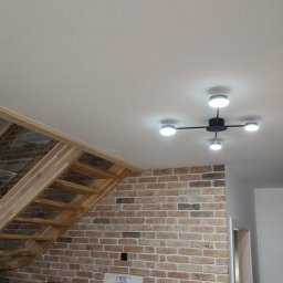 Mariusz Skonieczny - Nowoczesny żyrandol sufitowy z czterema okrągłymi lampami LED, zamontowany w jasnym wnętrzu z ceglaną ścianą i drewnianymi schodami z elementami szkła.