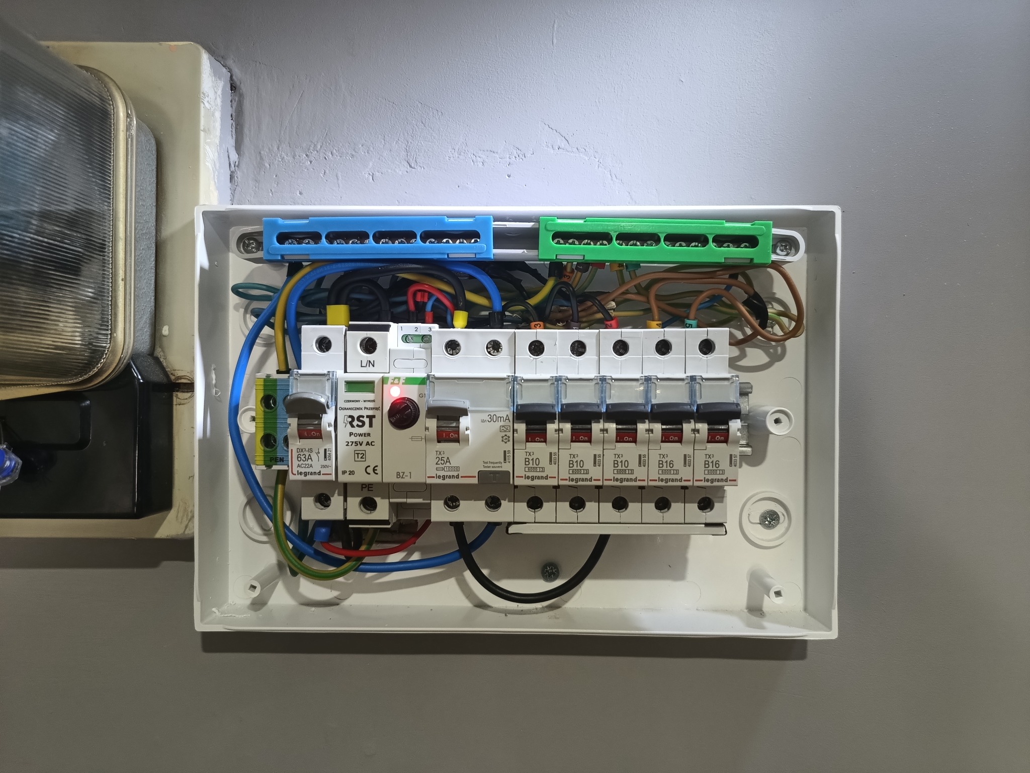 Rozdzielnia elektryczna z bezpiecznikami i okablowaniem w kolorach: niebieskim, zielonym, żółtym i brązowym. Widoczne oznaczenia i elementy marki Legrand.