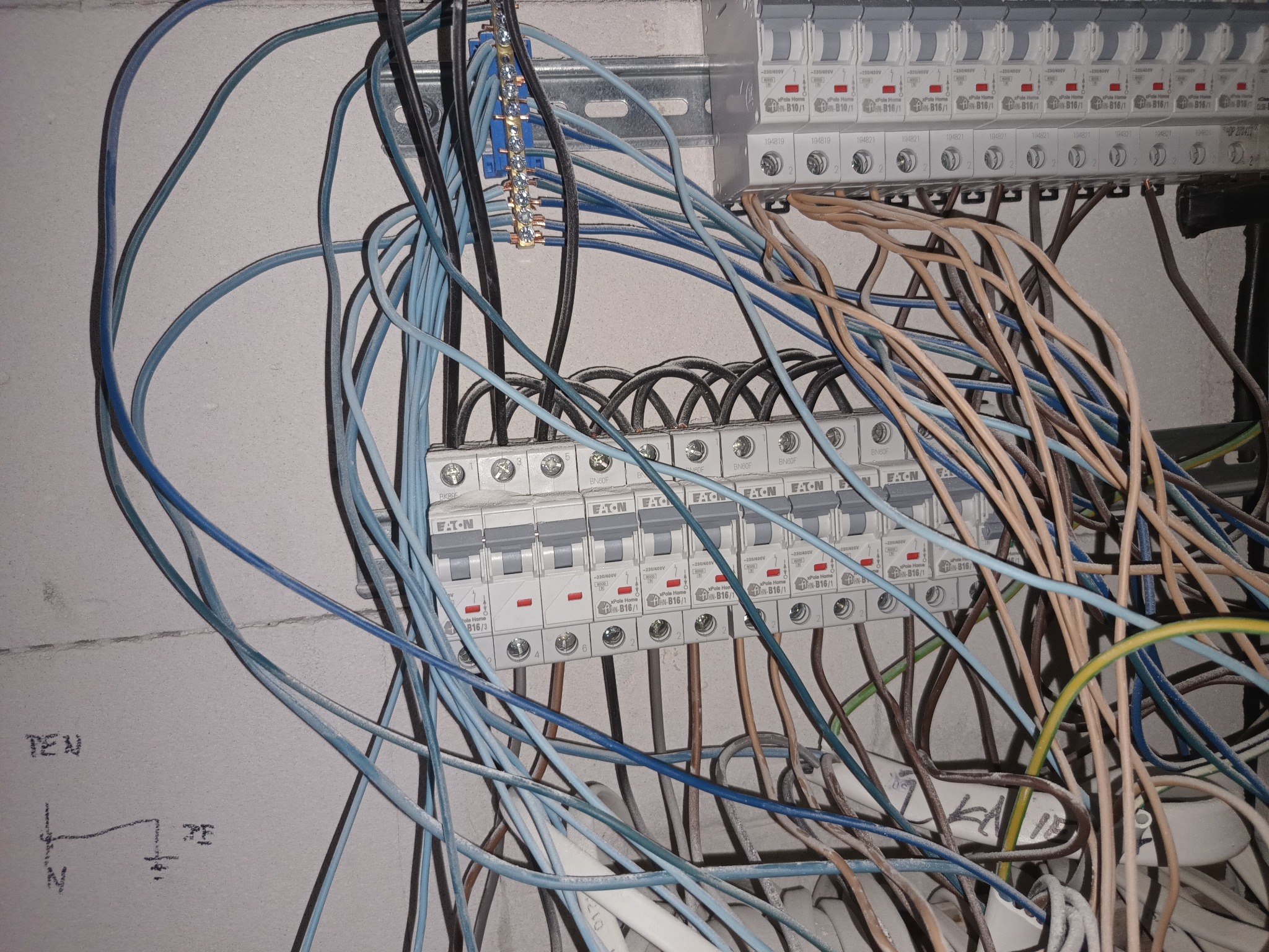 Rozdzielnica elektryczna z zamontowanymi bezpiecznikami EATON, widoczne liczne przewody w kolorach niebieskim, brązowym i czarnym,  schemat instalacji na ścianie.