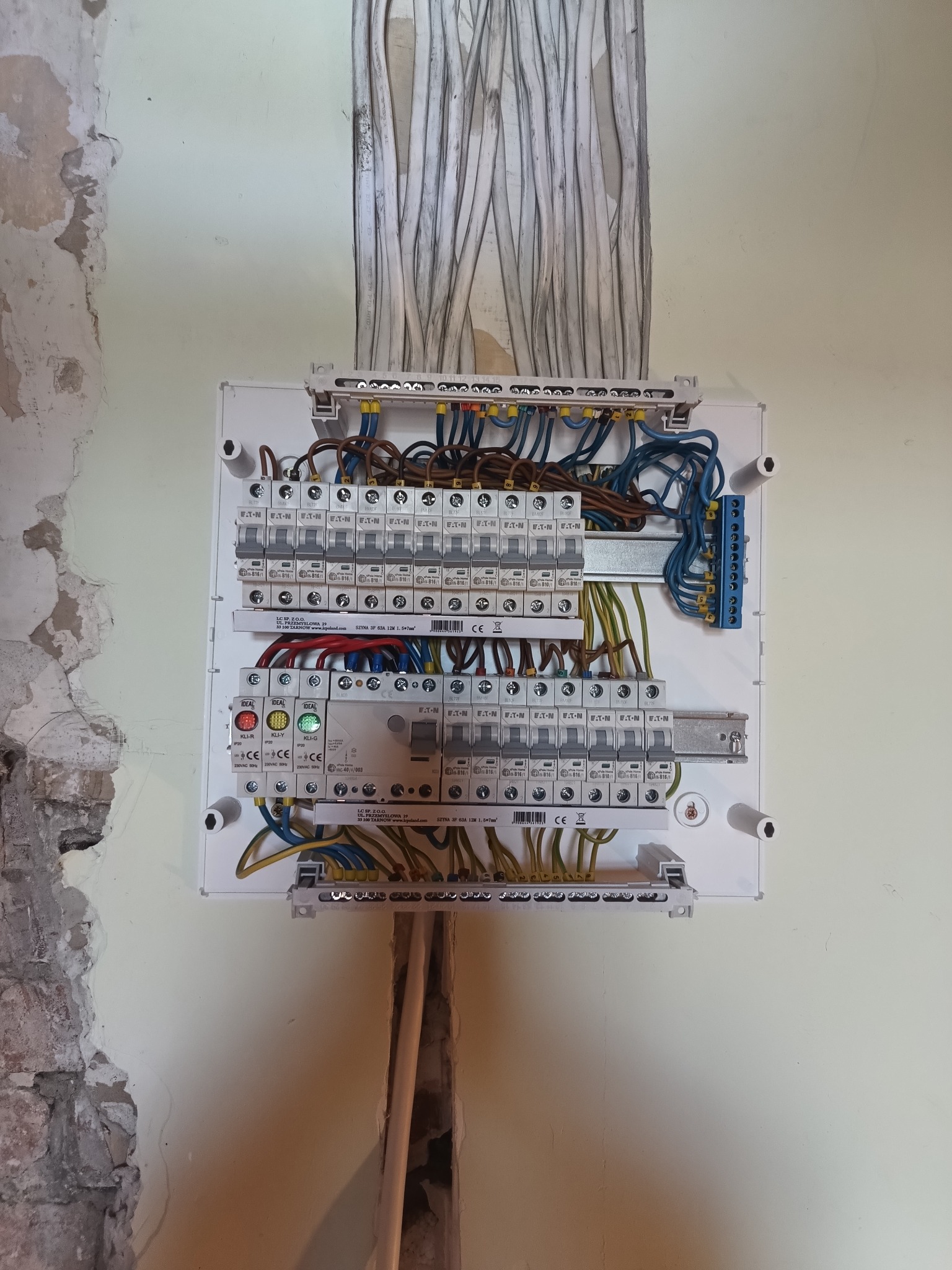Rozdzielnica elektryczna z licznymi bezpiecznikami i przewodami w trakcie montażu na ścianie z widocznymi śladami prac remontowych.