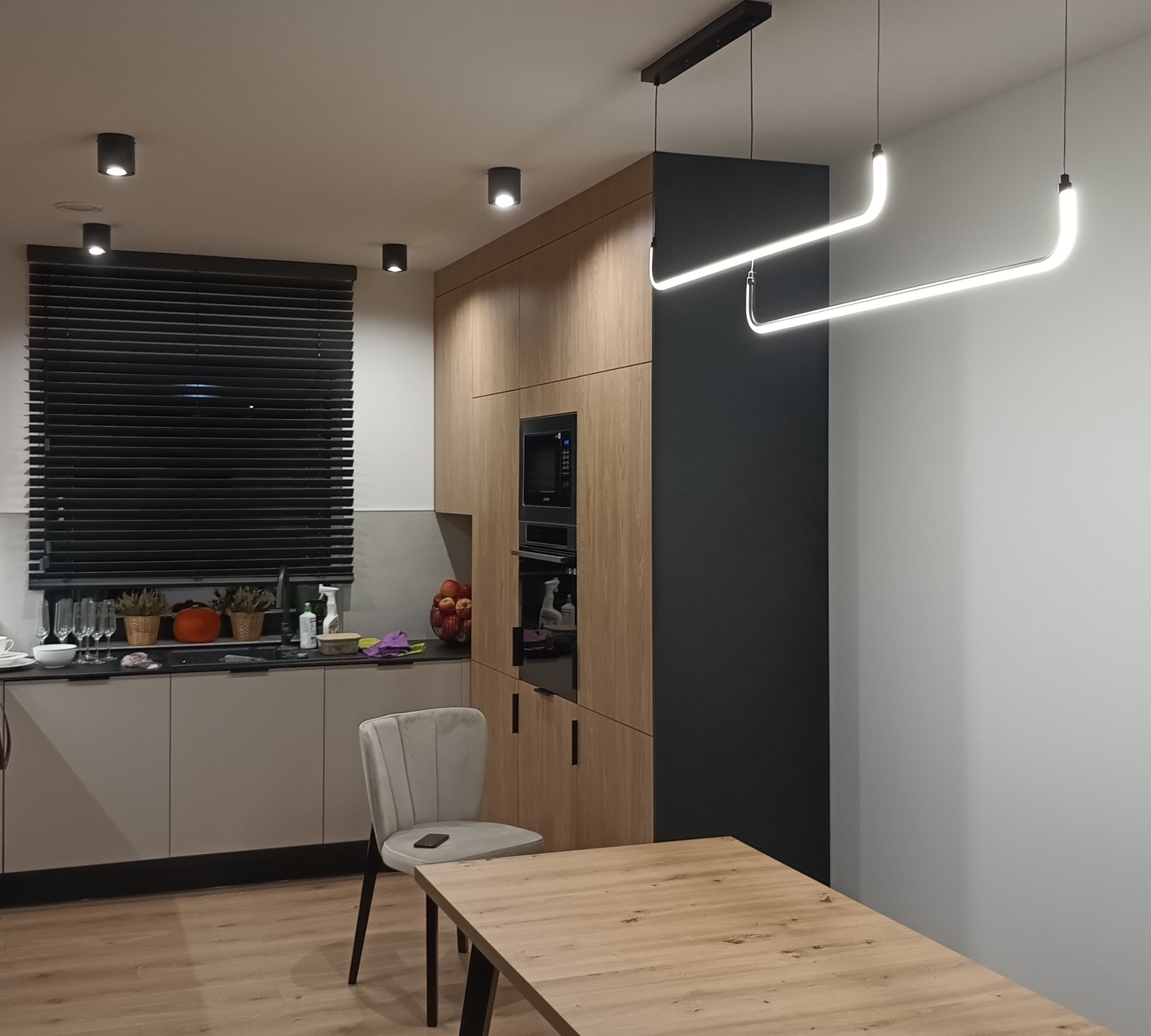 Nowoczesna kuchnia z drewnianą zabudową i designerskim oświetleniem LED. Widoczne lampy wiszące o unikalnym kształcie, punktowe światła sufitowe i żaluzje w oknie.