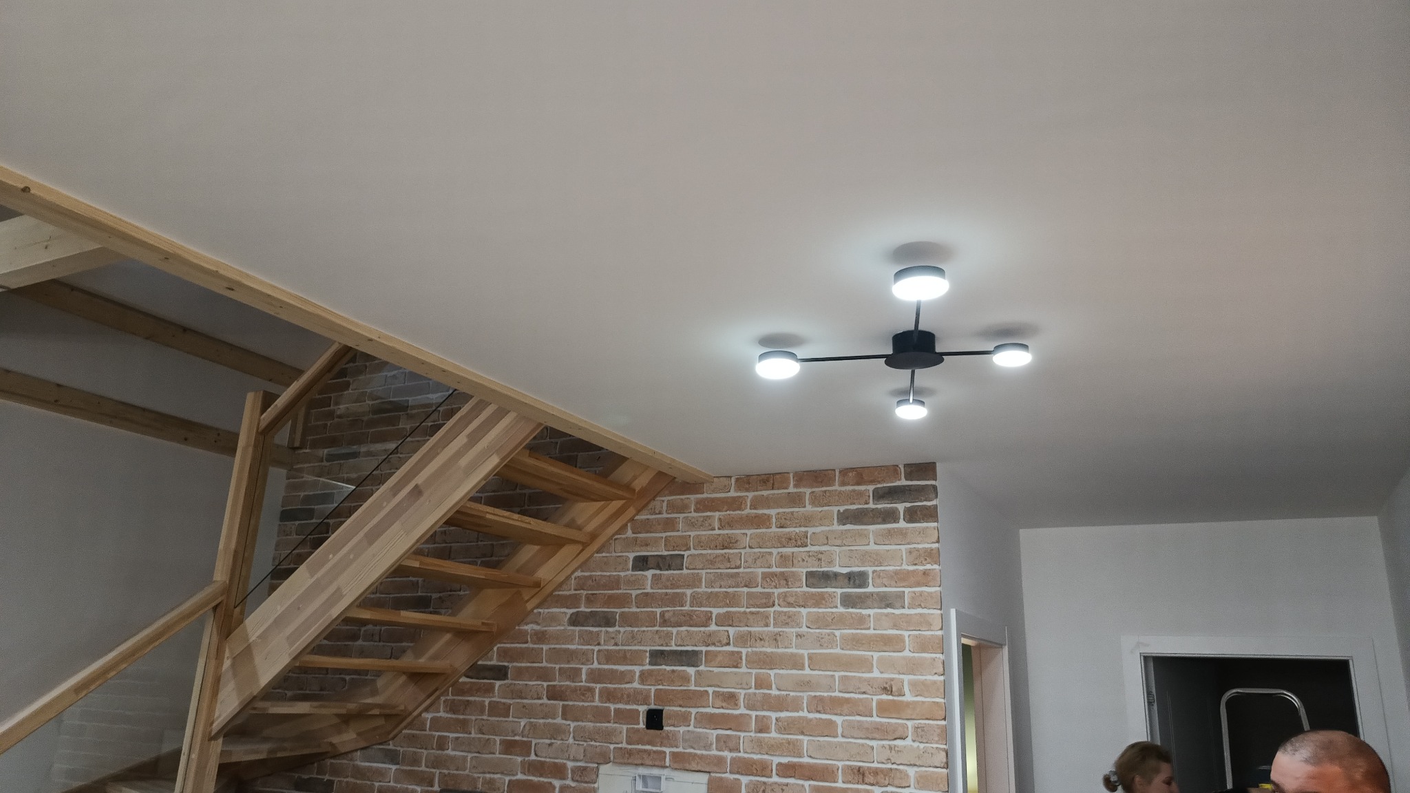 Nowoczesny żyrandol sufitowy z czterema okrągłymi lampami LED, zamontowany w jasnym wnętrzu z ceglaną ścianą i drewnianymi schodami z elementami szkła.